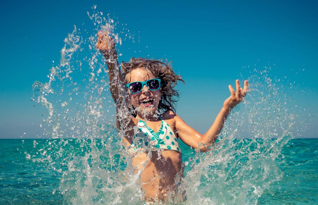 Bambina felice spruzza acqua al mare, cielo azzurro.